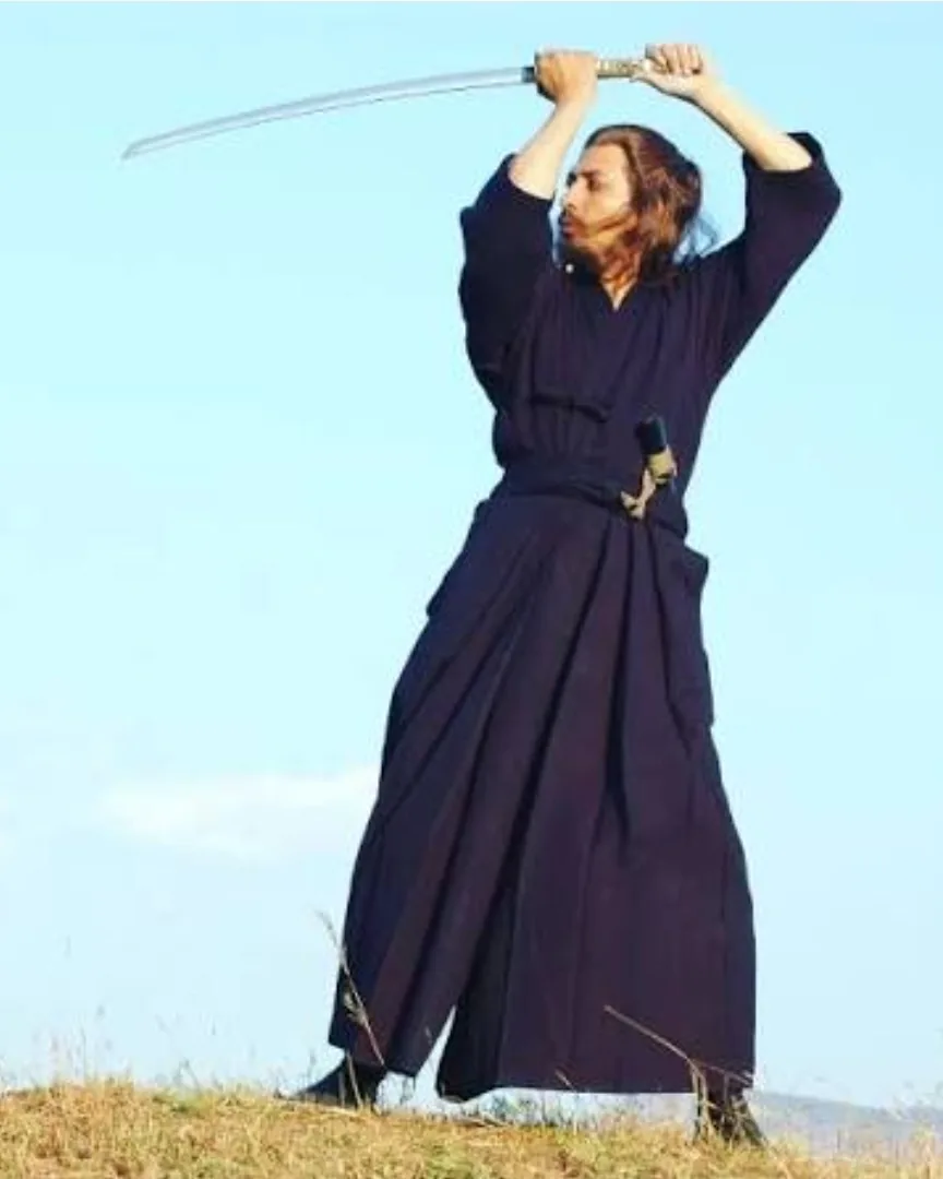 Swordsman in a navy keikogi and hakama raising a katana overhead on a hilltop